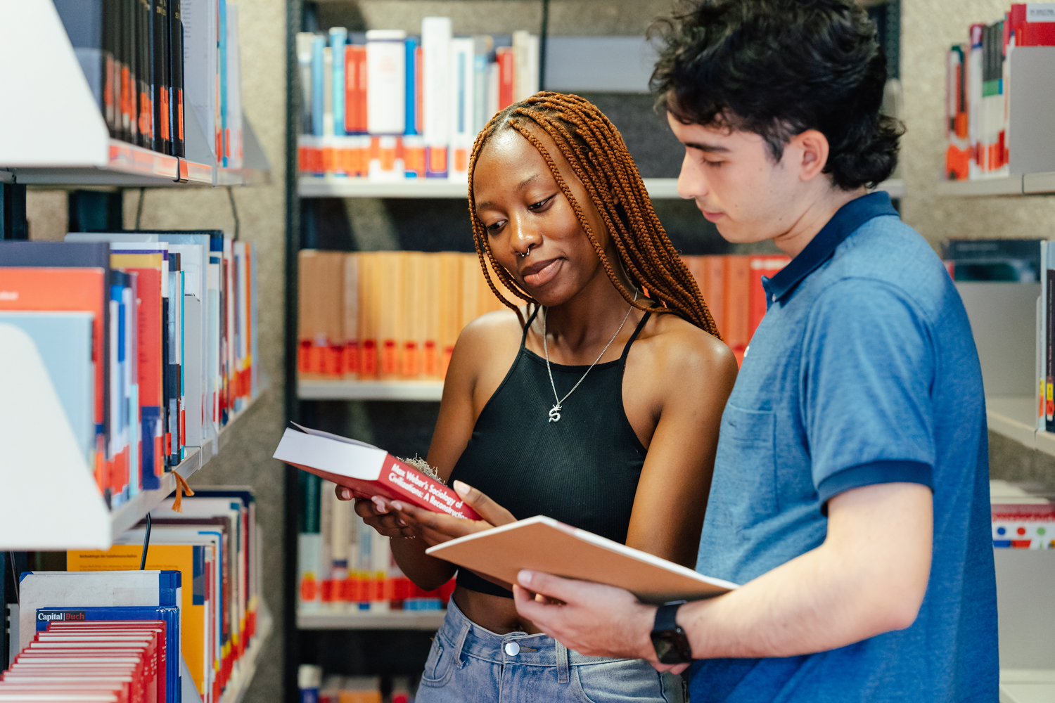 Two students stand in the university library holding textbooks.