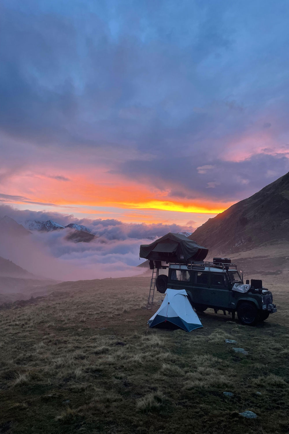 Ein Blick auf den Sonnenuntergang in den Bergen mit einem Auto mit Dachzelt und Campingzelten im Vordergrund.