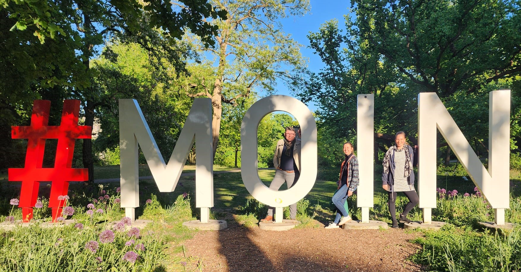 Zu sehen sind Henning Karsten, Maike Stelter und Melanie Schaller (Team Alumni) vor einem übergroßen Schriftzug 'Moin' in einem Stadtpark von Bremen.