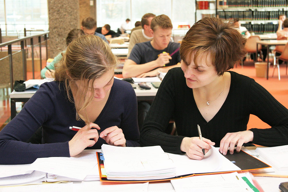 Zwei Studentinnen sitzen zusammen in der Bibliothek und schauen in ein Heft 