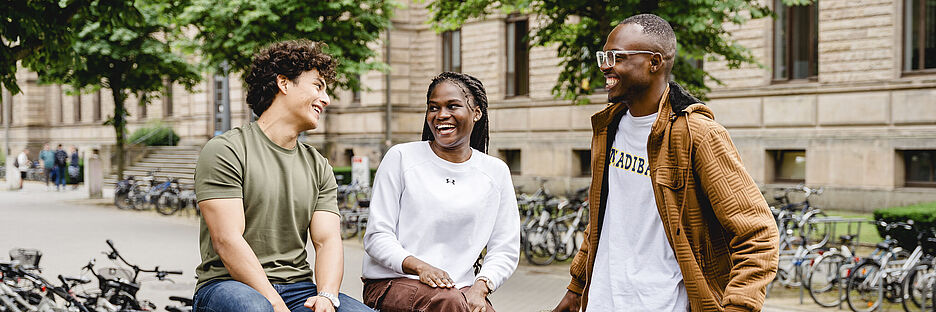 Three students sit on campus and talk to each other. The TU Braunschweig's Old Building can be seen in the background.