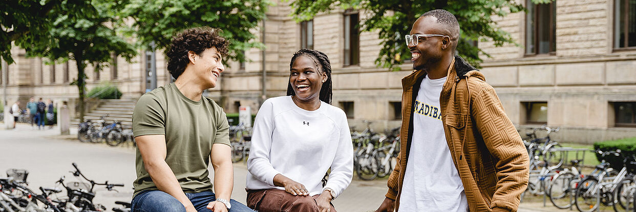 Drei Studierende sitzen auf dem Campus und reden miteinander. Im Hintergrund ist das Altgebäude der TU Braunschweig zu sehen. 