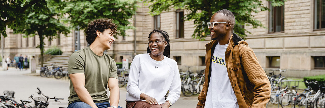 Drei Studierende sitzen auf dem Campus und reden miteinander. Im Hintergrund ist das Altgebäude der TU Braunschweig zu sehen. 