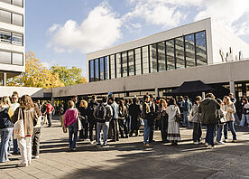 [Translate to English:] Many students are standing in groups on the university square in front of the main lecture hall.
