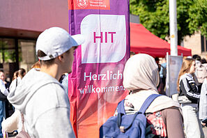 Schülerinnen und Schüler stehen vor einer Fahne, auf der das HIT Logo zu sehen ist und auf der herzlich Willkommen steht.