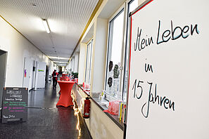 Das Bild zeigt einen winterlich geschmückten Flur, linksseitig steht eine Tafel, auf der interaktive Vorträge beworben werden. Rechts stehen festliche, rot bezogene Stehtische. Am rechten Bildrand im Vordergrund steht eine Planwand, auf der mein Leben in 15 Jahren geschrieben ist. Eine Person geht fokussiert den Gang hinunter.