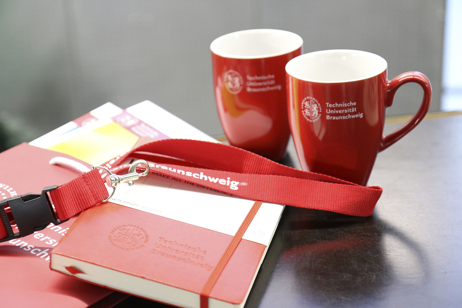 Zwei Tassen, eine Mappe, ein Notizbuch und ein Schlüsselanhänger im TU Design mit TU Braunschweig Logo