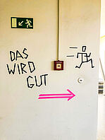 Auf einer hellen Wand klebt in schwarz der Schriftzug „DAS WIRD GUT“. Rechts davon ein stilisiertes Strichmännchen aus schwarzem Klebeband mit Bewegungsstrichen, das nach rechts eilt. Unter all dem ein pinker Pfeil, der nach rechts weist.