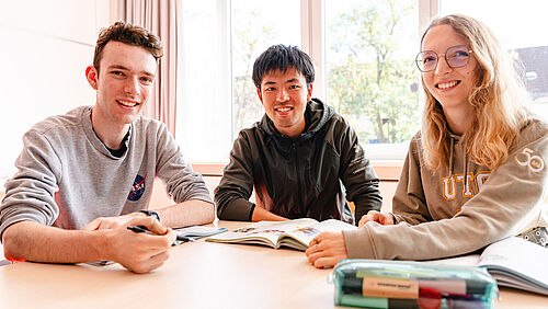 Drei Studierende sitzen in einem Unterrichtsraum und lächeln in die Kamera. Auf dem Tisch liegen Lehrbücher.