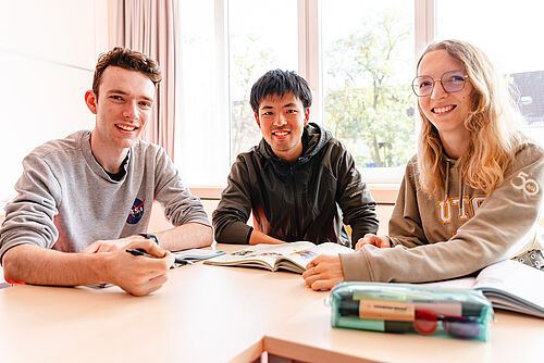 Drei Studierende sitzen in einem Unterrichtsraum und lächeln in die Kamera. Auf dem Tisch liegen Lehrbücher.