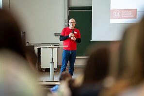 Ein Mitarbeiter der TU Braunschweig steht in einem Hörsaal vor einer Tafel. Er hält ein Mikrofon in der Hand. Im Vordergrund sind junge Menschen zu erkennen
