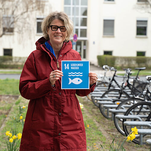 Prof. Dr. ès sci. Antje Schwalb steht auf dem Campus und hält das Schild zum 14. SDG. 