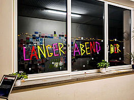 Innenaufnahme eines Flurs bei Dunkelheit mit drei nebeneinanderliegenden Fenstern. Auf den Scheiben kleben in leuchtenden Neonfarben (Pink, Rot, Gelb, Grün) aus Klebeband die Großbuchstaben „LANGER ABEND DER“. Um die Buchstaben verteilen sich weiße Schneeflocken-Aufkleber. Durch die Scheiben spiegelt sich eine gegenüberliegende Wand, die dicht mit bunten Zetteln und Postkarten sowie einer warm leuchtenden Lichterkette dekoriert ist.