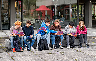 Studieninteressierte sitzen auf dem Universitätsplatz und stöbern durch HIT-Programmhefte