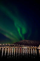 Polarlichter in Finnland. Im Vordergrund ist eine beleuchtete Brücke, im Hintergrund ein Bergpanorama zu sehen.