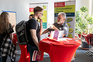 Zwei Männer stehen an einem roten Stehtisch, auf dem Taschen mit dem TU Logo und der Aufschrift willkommen liegen. Auf dem Stehtisch steht ein Schild auf dem steht Immatrikulationsamt. Im Hitnergrund sieht man zwei Roll Ups, auf dem einen steht Studienservice Center.