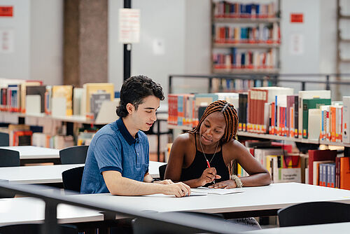 Zwei Studierende sitzen in der Universitätsbibliothek an einem Tisch und lernern.