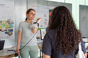 Zwei Personen unterhalten sich an einem Messestand. Eine Person hat eine VR-Brille in der Hand