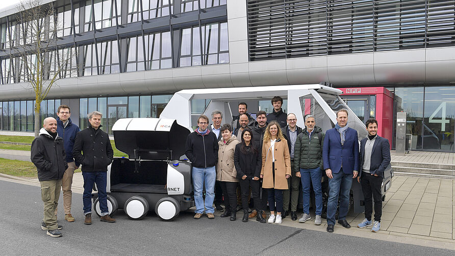 Abschlussveranstaltung im LogiSmile-Projekt am 22. März 2024 am NFF: Gruppenfoto Projektmitglieder, im Hintergrund die beiden Forschungsfahrzeuge RAION und ONA