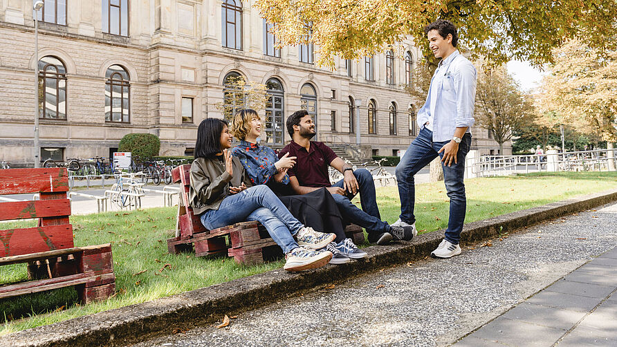 Three international students are sitting on a pallet bench. Another student is standing next to them. They are all chatting. The old TU Braunschweig building can be seen in the background.