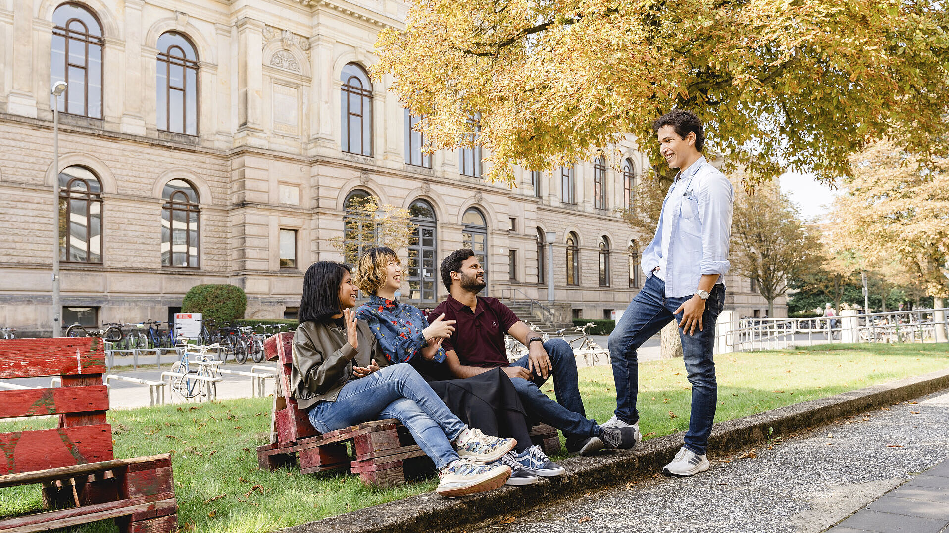 Three international students are sitting on a pallet bench. Another student is standing next to them. They are all chatting. The old TU Braunschweig building can be seen in the background.