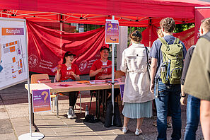 Zwei Menschen sitzen an einem Tisch unter einem roten Pavillon, lachen und geben Hefte aus. Vor ihnen steht eine Schlange an Menschen und wartet