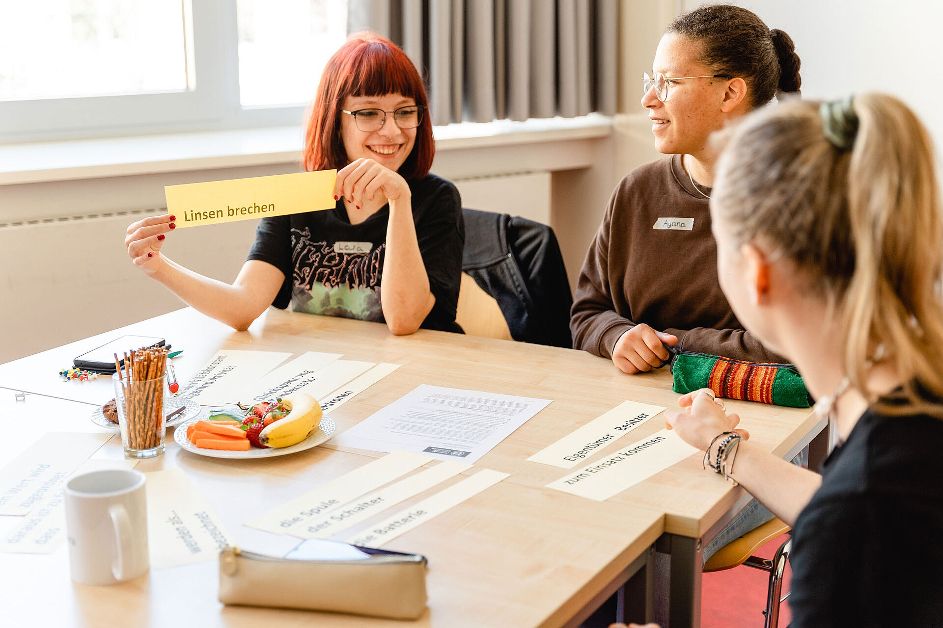 Drei Studiernde sitzen an einem Tisch. Eine von ihnen hält ein Schild mit dem Fachbegriff "Linsen brechen" in die Höhe.