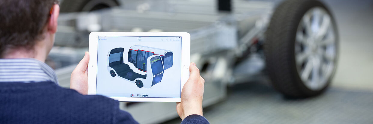 Wissenschaftlicher hält ein Tablet mit einer Vision einer autonomen Fahrzeug in der Hand. Im Hintergrund: die Fahrzeugplattform.  