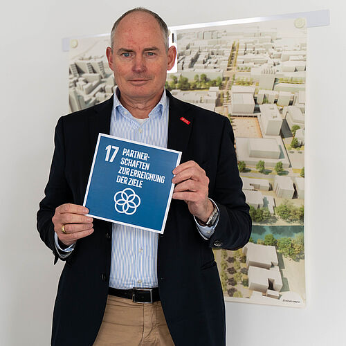 Dietmar Smyrek hält eine Karte in beiden Händen. Darauf ist das 17. SDG zu sehen. Er steht in seinem Büro.