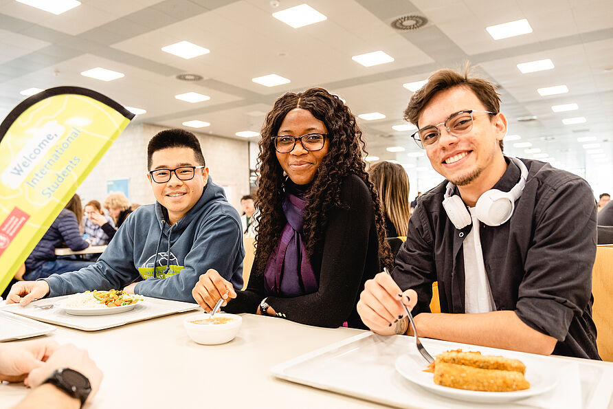 Drei internationale Studierende sitzen in der Mensa und nehmen an der International Lunch Break teil.