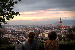 Blick von einem Aussichtspunkt über eine italienische Stadt in der Dämerung. Im Vordergrund sind Menschen zu sehen, die den Ausblick genießen.