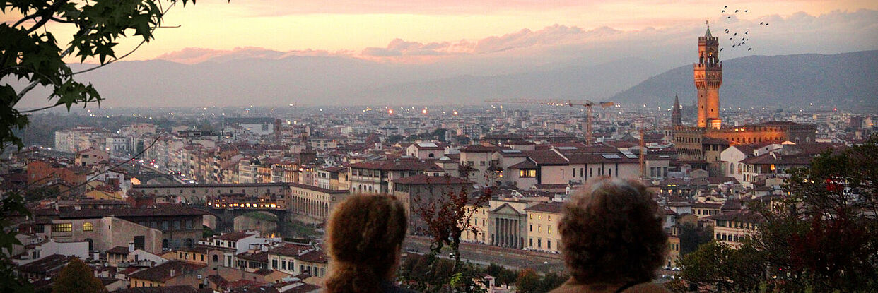 Blick von einem Aussichtspunkt über eine italienische Stadt in der Dämerung. Im Vordergrund sind Menschen zu sehen, die den Ausblick genießen. 