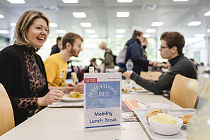 Mehrere Personen sitzen an einem Tisch in der Mensa der TU Braunschweig und reden miteinander. Auf dem Tisch steht ein Aufsteller mit der Aufschrift "Mobility Lunch Break". 