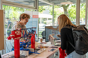 Ein Mitarbeitender der TU Braunschweig unterhält sich an einem Info-Stand mit einer Schülerin. Auf dem Tisch steht ein Monitor und ein Gebilde aus Lego.