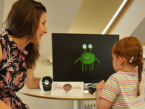 Katie Von Holzen und Kind vor ein Bildschirm mit Alien darauf, im Vordergrund ist der Ini-Seg Logo