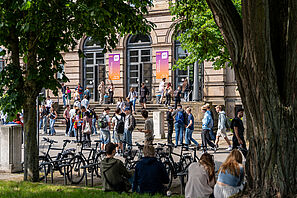Man sieht eine Menschenmenge vor dem Altgebäude der TU, an den Seiten sieht man Bäume und grünen Rasen. Auf dem Rasen sitzen einige Menschen. An der Fassade des Altgebäudes hängen 2 Banner mit dem HIT Logo. Auf ihnen steht dein Tag an der Uni