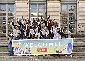  A large group of international students stands in front of the old building holding a banner that reads ‘Welcome’.
