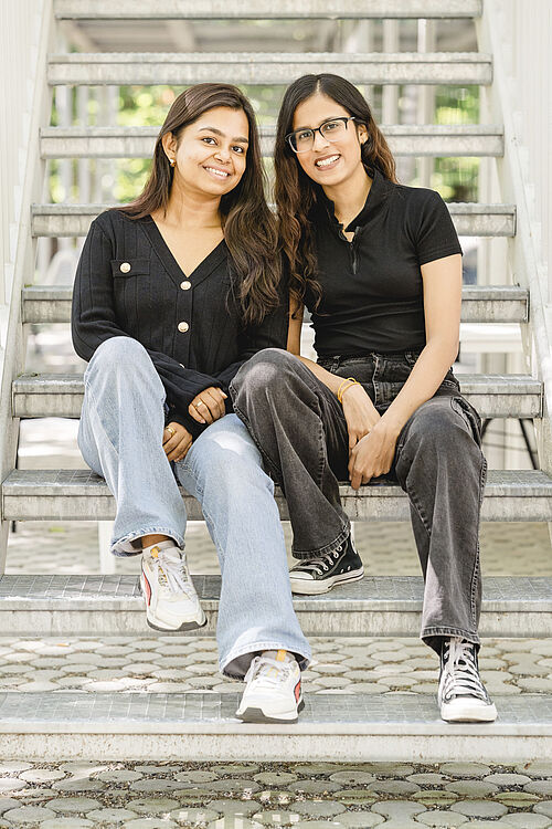  Two female students, who are members of the Indian Student Community at TU Braunschweig, are sitting on the steps in front of the student union building.