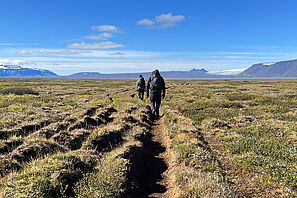 Zwei Studierende wandern über eine Hochebene in Island. Im Hintergrund ist ein schneebedecktes Bergpanorama. 