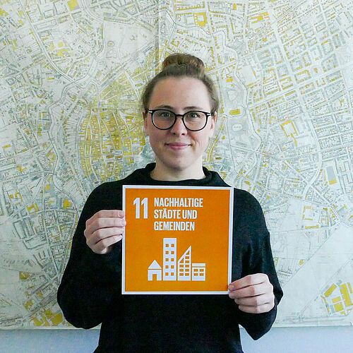 Larisa Tsvetkova hält die SDG 11 Karte und steht vor einem Stadtplan. 