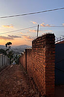 Blick auf ein Tal, in dem die Stadt Kigali zu sehen ist, bei Sonnenuntergang. Im Vordergrund ist ein schmaler, von Mauern flankierter Weg zu sehen.