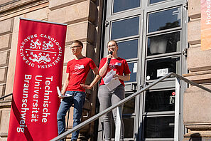 Zwei Studierende in roten TU-Braunschweig T-Shirts stehen auf der Treppe des Altgebäudes neben einem TU Roll-Up. Sie halten Mikrofone und Zettel in der Hand und lächeln. 