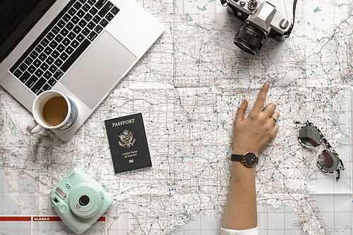 Eine Person studiert eine Karte, auf der ein Laptop, eine Kaffeetasse, zwei Kameras, ein amerikanischer Pass und eine Sonnenbrille liegen.