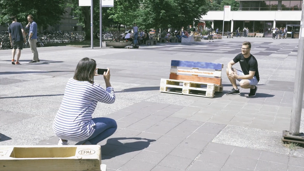 A woman takes a photo of a man next to a bench made of Euro pallets in the Forum Square