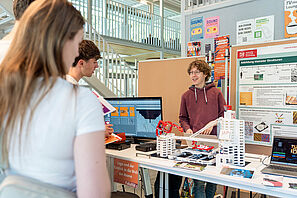Ein freundlich lächelnder Mann steht an einem Messestand, auf dem Lego und ein Monitor aufgebaut ist. Im Vordergrund stehen interessierte Menschen