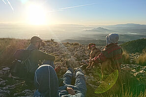 Drei Studierende sitzen mit einem Hund auf einem Berg und genießen die Aussicht über die spanische Berglandschaft.