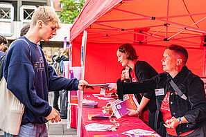 Ein Studieninteressierter steht vor einem roten Zelt und erhält ein Programmheft