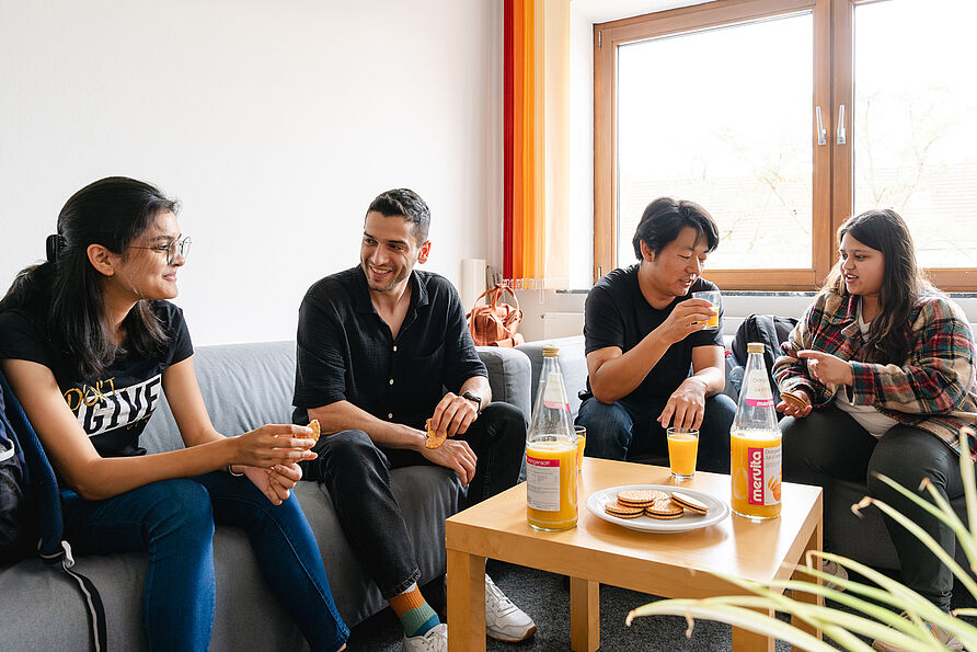 Vier Studierende sitzen auf Sofas und unterhalten sich miteinander. Auf dem Tisch stehen Kekse und Getränke.