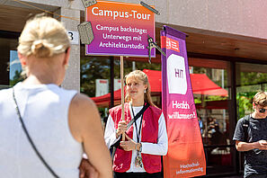 Person mit roter Weste hält ein Schild mit der Aufschrift 'Campus-Tour Campus backstage: Unterwegs mit Architekturstudis' vor einem Gebäude mit einem Banner 'HIT Herzlich willkommen!'