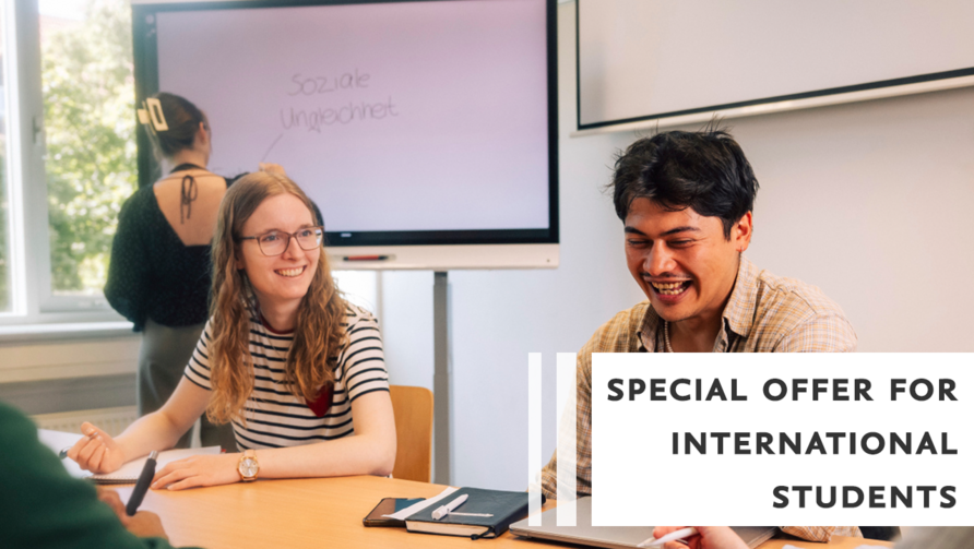 Studierende sitzen an einem Tisch im Seminarraum und tauschen sich aus. Im Hintergrund schreibt jemand an ein Whiteboard.